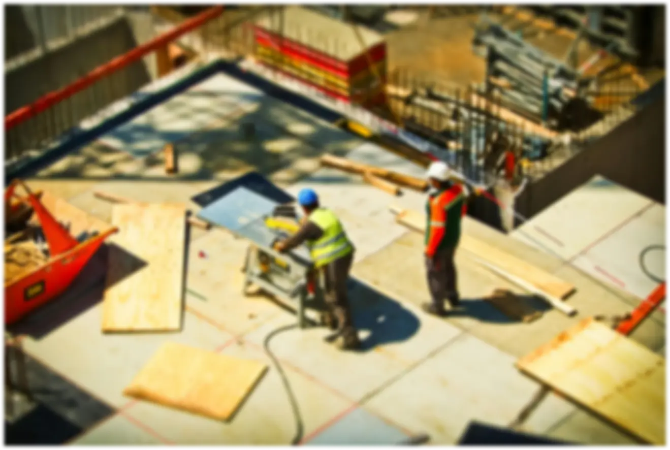 Poză blurată cu un șantier de construcții, în care sunt prezenți doi muncitori, lucrând.
